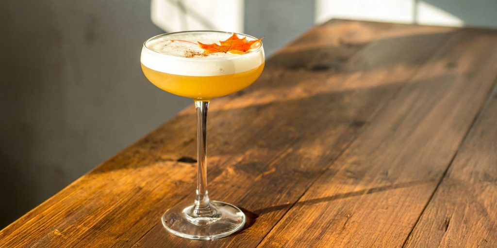 Maple Whiskey Sour cocktail in a coupe glass, garnished with an orange peel, surrounded by vibrant orange maple leaves on a white surface.