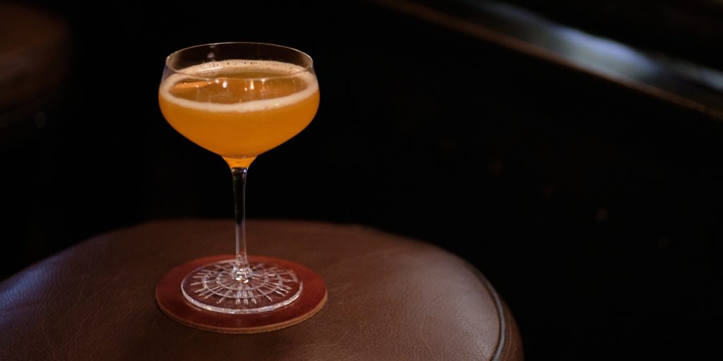 Mules Hind Leg cocktail in a coupe glass with a frothy top, served on a metal tray with a jigger, alongside scattered maple leaves. Image by Mykola Khalkha on Adobe Stock.