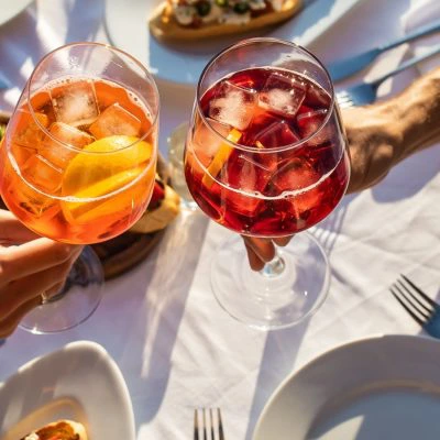 Clinking 2 glasses, one with Campari and the other with Aperol at a table set with appetisers