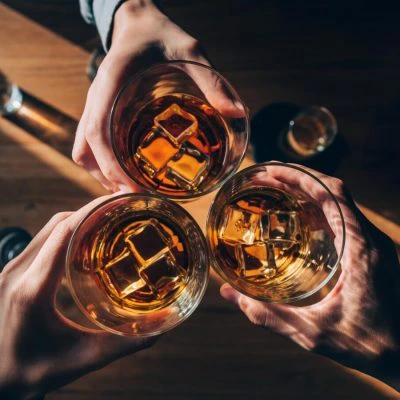 Three glasses of bourbon with ice being clinked over a table