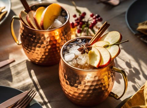 Copper mugs filled with a Thanksgiving batch cocktail of Apple and Cinnamon Mule, garnished with ice, fresh apple slices, cinnamon sticks, and star anise, set on a harvest-themed table with golden napkins and warm lighting.
