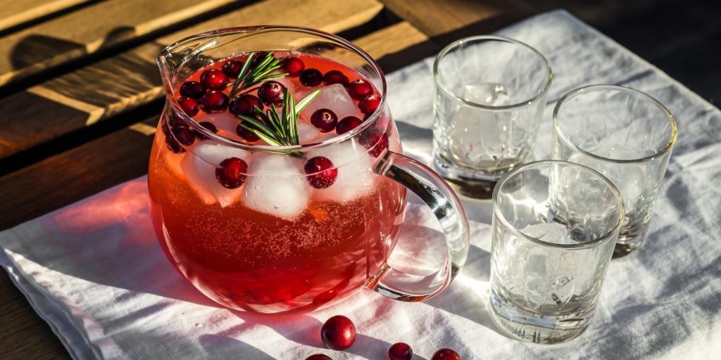 Thanksgiving batch cocktails like this Cranberry Vodka Spritz pitcher offer a visual feast: a clear glass jug filled with icy pink spritz, dotted with floating cranberries and sprigs of rosemary. It’s placed on a white cloth in soft autumn light, with empty glasses nearby—ready for guests. The cranberry-red hue evokes falling maple leaves, echoing the season’s festive, shareable spirit