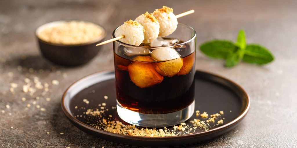 A Hong Kong–inspired whiskey cocktail with black sesame, perfect for Thanksgiving batch cocktails. Served in a short glass with large ice cubes and garnished with toasted rice balls on a skewer. Crushed sesame and a sprig of mint decorate the plate.