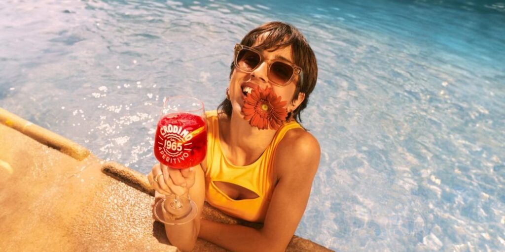 A young lady with sunglasses on a a flower in her teeth standing in a pool with a glass of non-alcholic Rosso Crodino.