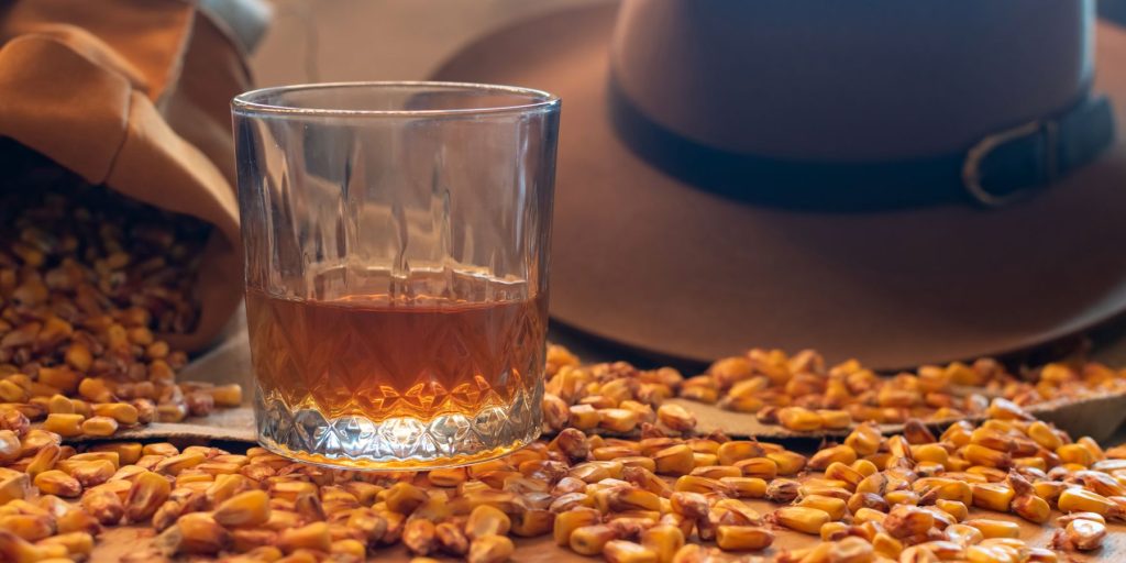 A glass of bourbon surrounded by corn with a hat in the background