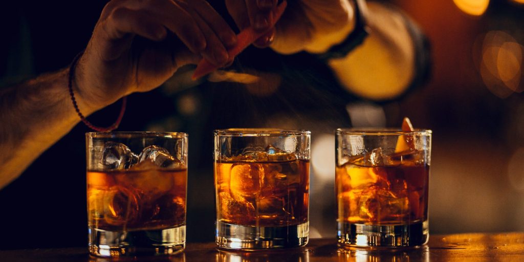 Three bourbon cocktails on a bar with a bartender squeezing and orange peel over the cocktails