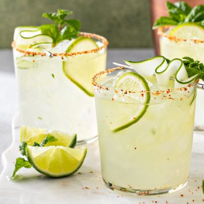 Three Margarita cocktails garnished with limes and cucumber ribbons on a marble surface.