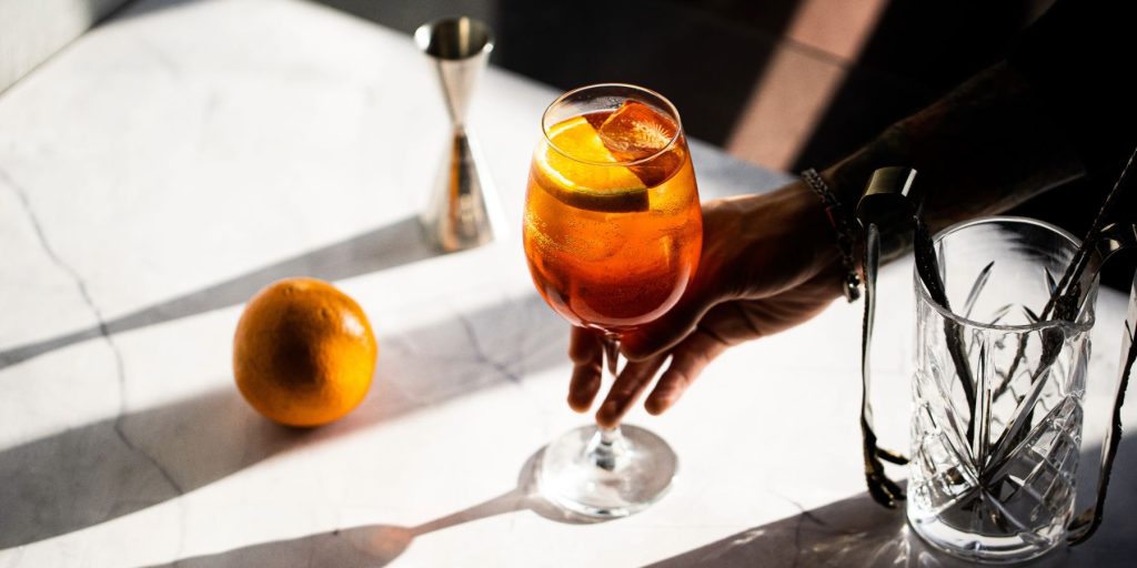 A hand holding an Aperol spritz cocktail on a white marble surface surrounded by cocktail tools and a fresh orange.