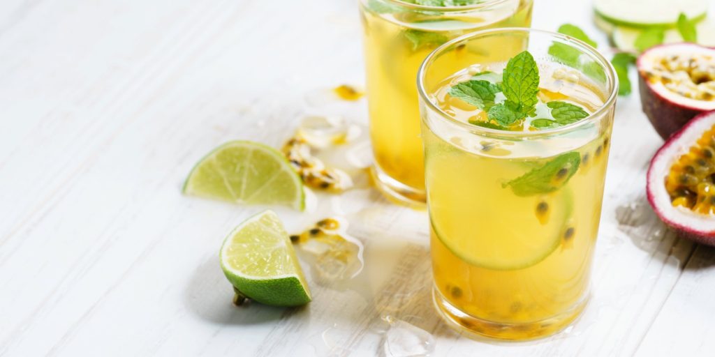 Two passion fruit cocktails garnished with mint, fresh limes and passion fruit on a white wooden surface.