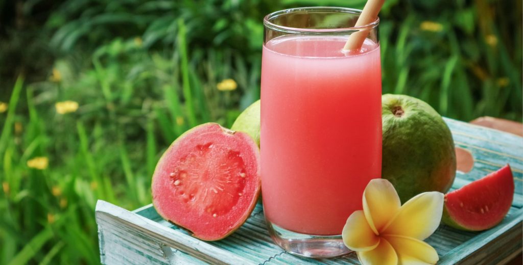 A guava cocktail on a blue wood tray surrounded by whole and cut guavas with a flower.