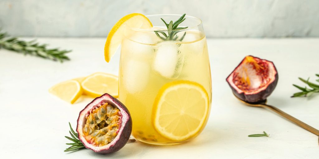 A passion fruit cocktail garnished with fresh lemon and rosemary on a white surface with fresh passion fruit halves.