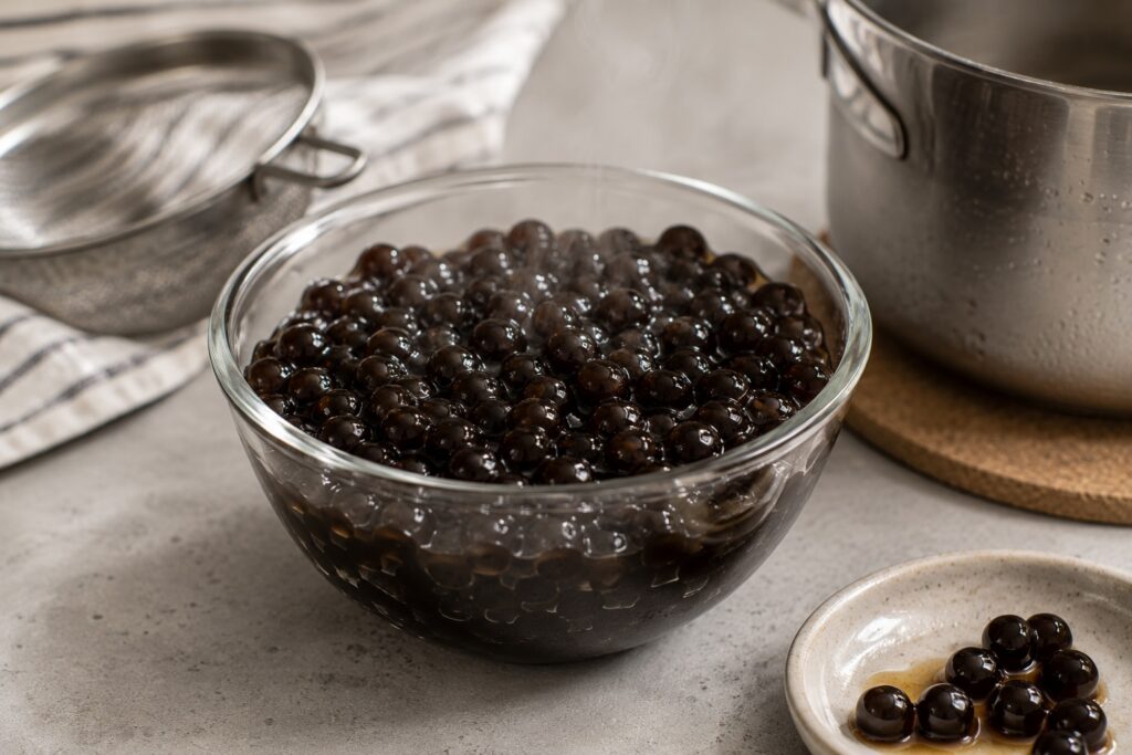 A glass bowl filled with freshly cooked tapioca pearls sits on a modern kitchen counter, with steam rising from the glossy black pearls. A saucepan, fine-mesh strainer, and small dish of pearls in syrup are in the background.