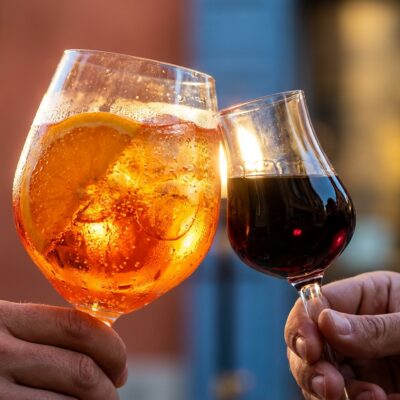 Two hands clinking an Aperol Spritz and an amaro digestif glass in explosive golden-hour backlight.