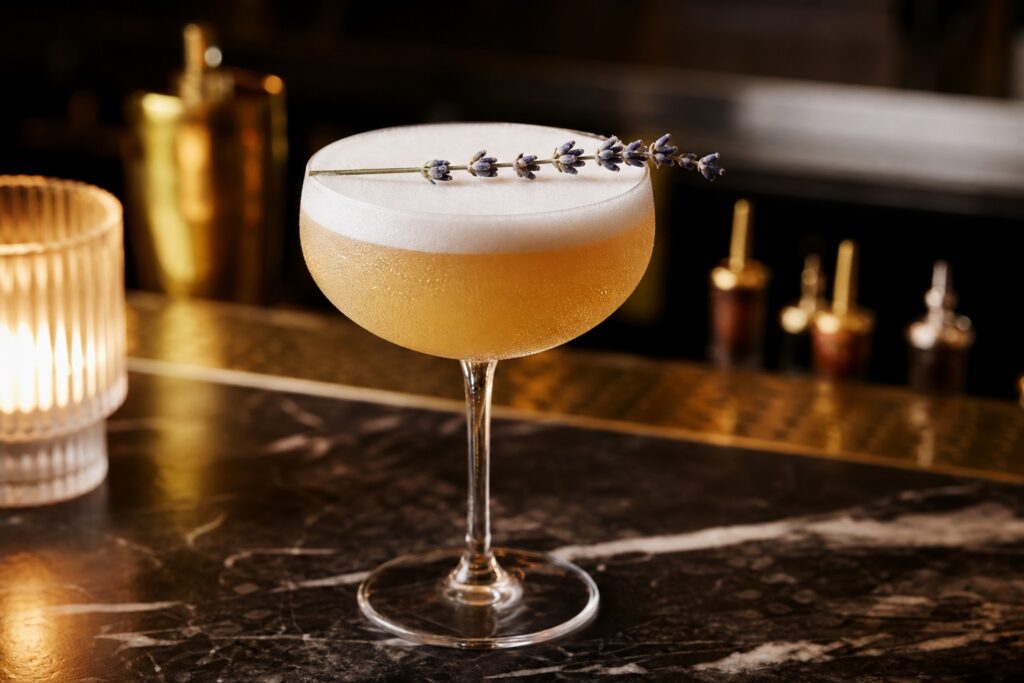 A gin sour cocktail in a chilled coupe glass, pale amber with a smooth, foamy top, garnished with a fresh lavender stem resting lightly on the foam surface, set against a softly blurred bar counter in warm, diffused light.