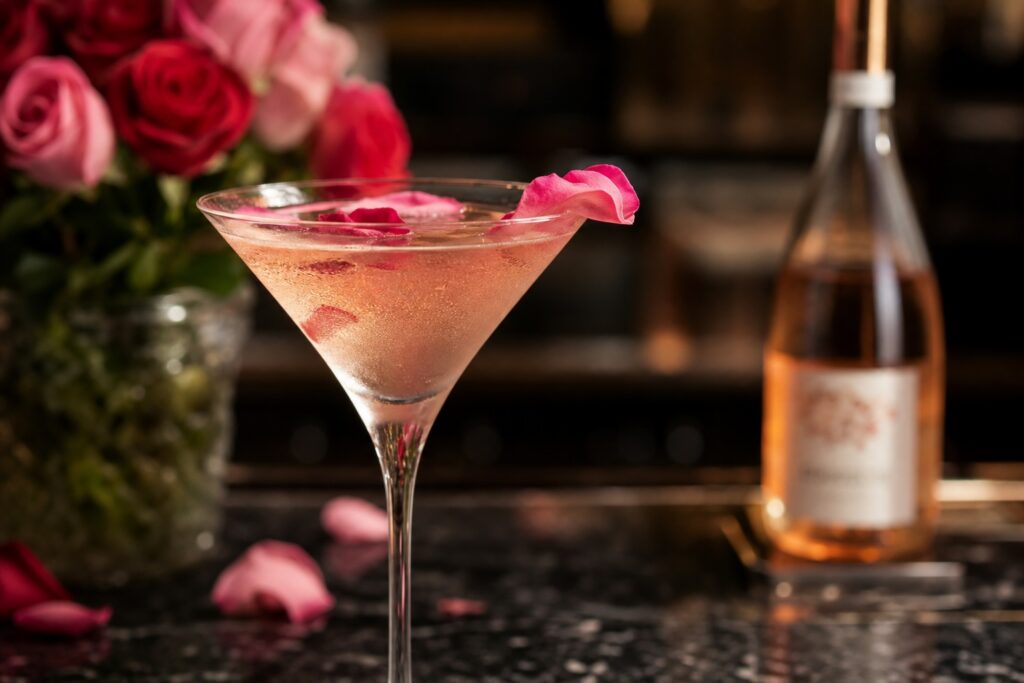A pale pink spritz in a martini glass, no ice, garnished with rose petals and a bottle of rosé wine and roses softly blurred in the background.