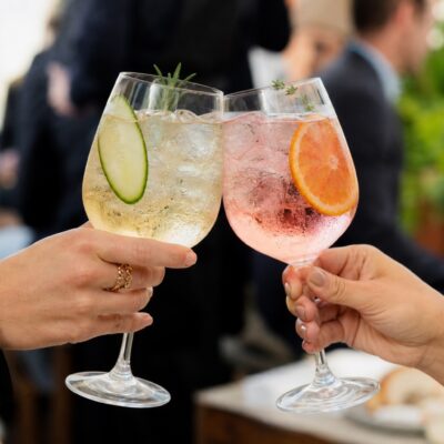 Two hands clink large gin and tonic glasses in a realistic close-up, with one drink pale gold and the other blush pink, both filled with ice and garnished for a celebratory toast.
