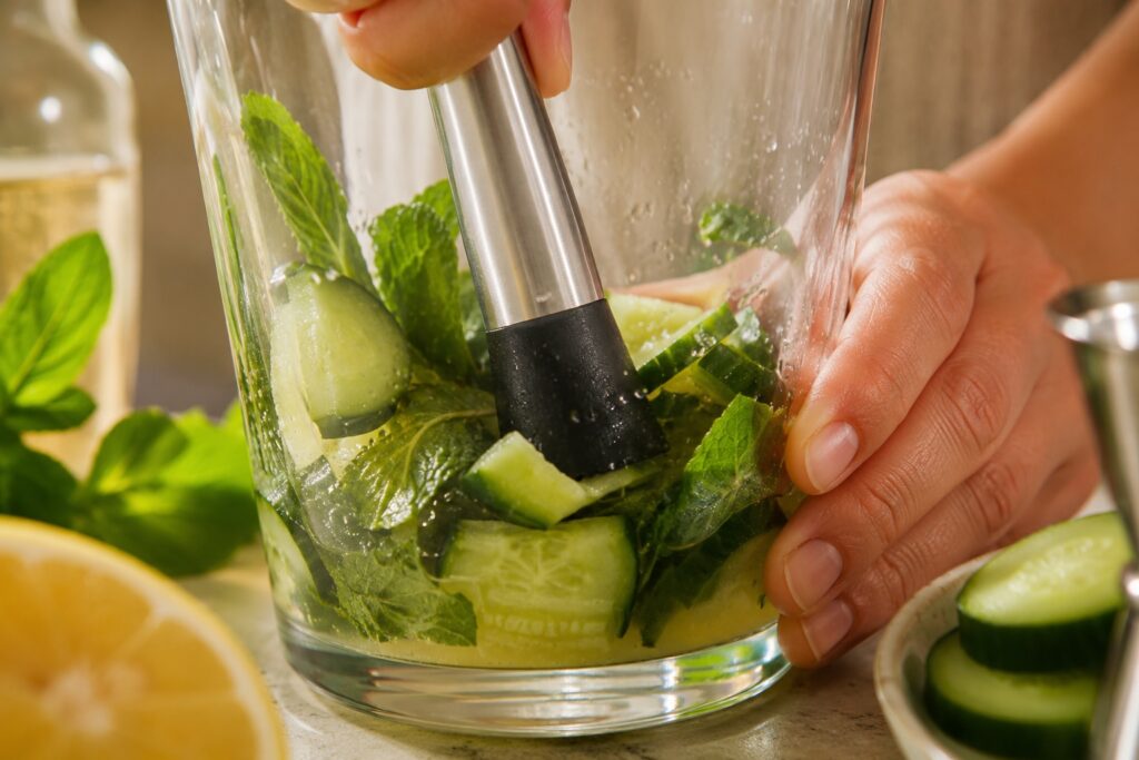Close-up of the preparation of a Cucumber Smash cocktail, with fresh cucumber, mint, lemon juice and simple syrup being gently muddled in a mixing glass before adding gin and ice.